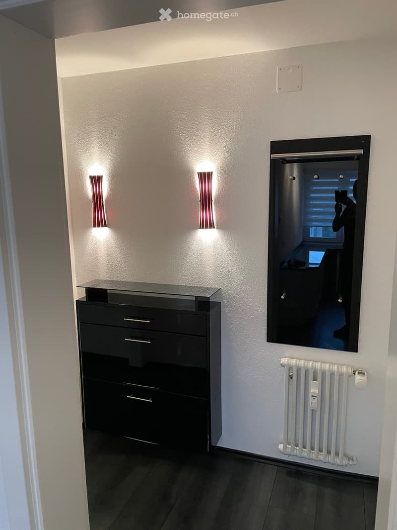 Modern hallway with sleek black cabinet, wall-mounted lights, radiator, and a large mirror reflecting a room beyond.