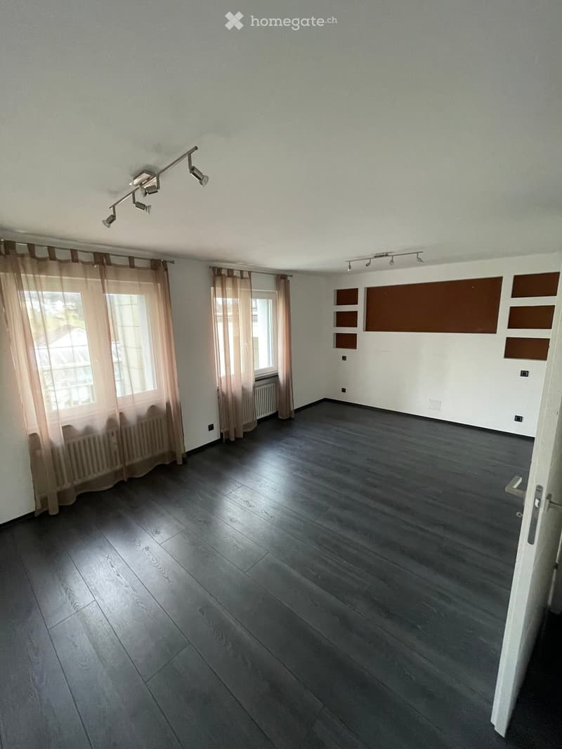 Empty room with dark wooden flooring, white walls, large windows with sheer curtains, and track lighting on the ceiling.