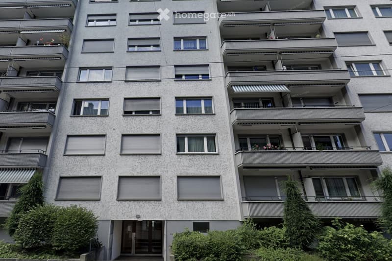 Multi-story apartment building with multiple balconies, large windows, and greenery around the base.