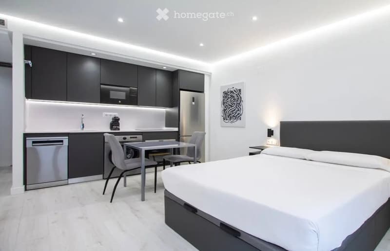 Modern studio apartment with a bed, small dining table, chairs, and a compact kitchen area featuring stainless steel appliances.