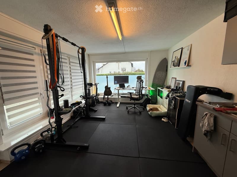 Well-lit room with exercise bike, treadmill, guitar, computer, chair, dog bed, surfboard, framed pictures, kettlebell, and headphones.