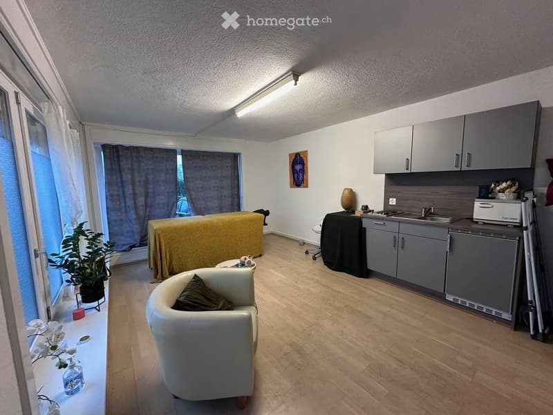 Modern furnished room with a sofa, small table, kitchen area with cabinets, sink, and a refrigerator. There is a massage table covered with a yellow sheet, a plant, and a picture on the wall.