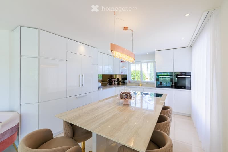 Modern kitchen with white cabinetry, a central island, and a cake on display. Features include a hanging light fixture, a sink, and an oven.