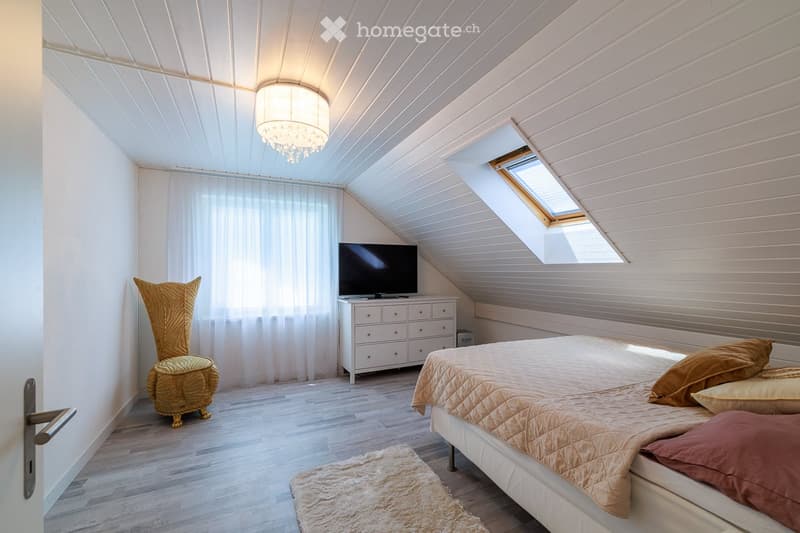 Cozy attic bedroom with wooden slat ceiling, skylight window, white walls, and modern furnishings including a bed, TV, dresser, and decorative chair.