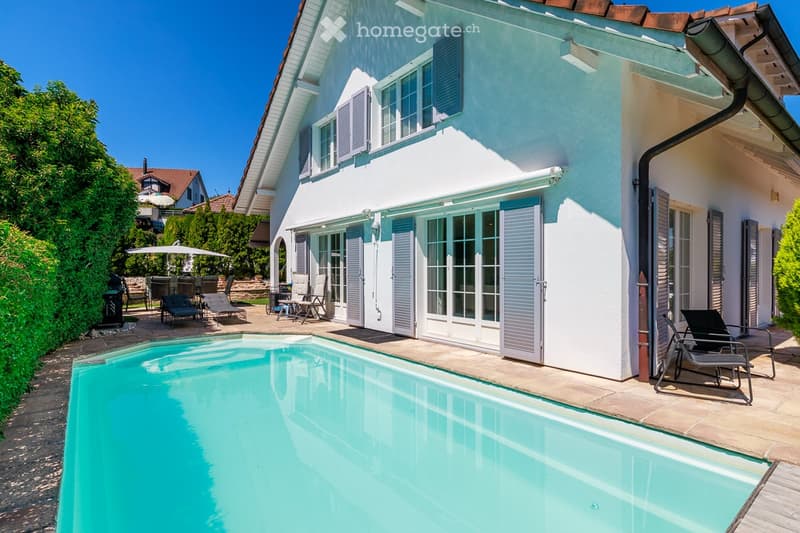 Two-story house with white exterior, blue shutters, large windows, and a swimming pool in the garden area.