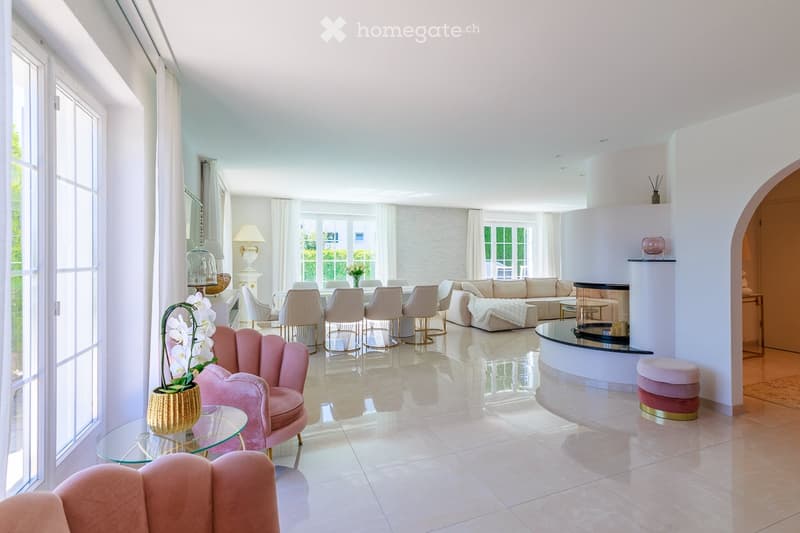 Bright, airy living room with large windows, white walls, and a mix of modern and classic furniture including a pink armchair, sofa, coffee table, and dining area with chairs.