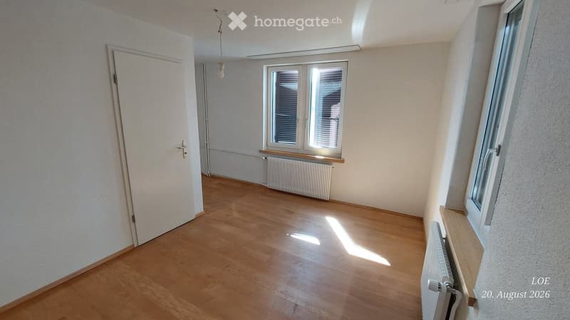 Empty room with wooden flooring, white walls, and large windows allowing natural light. Features include a radiator and a closed door.