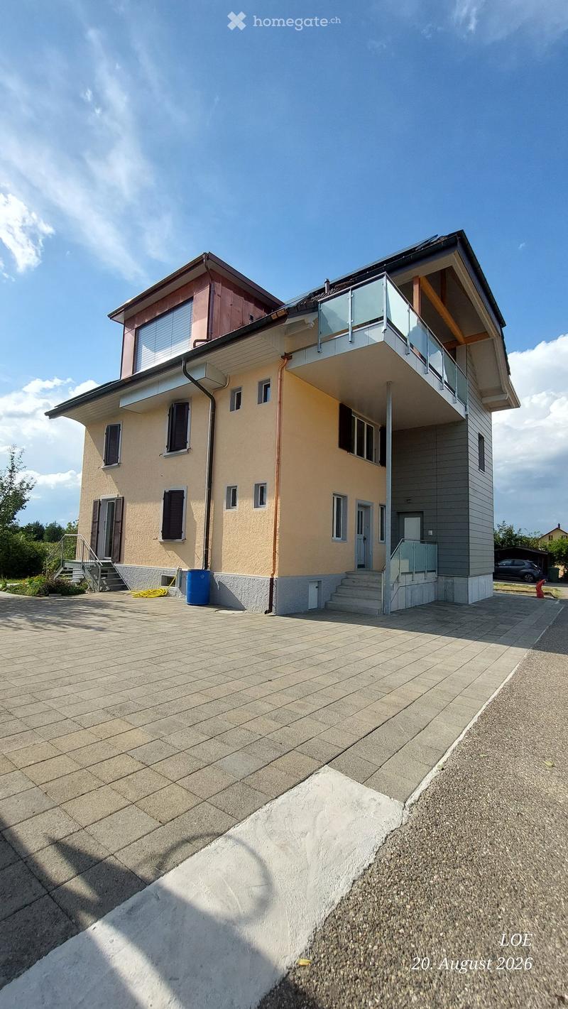 Two-story house with beige and grey exterior, multiple windows, a balcony on the upper level, and a paved driveway.