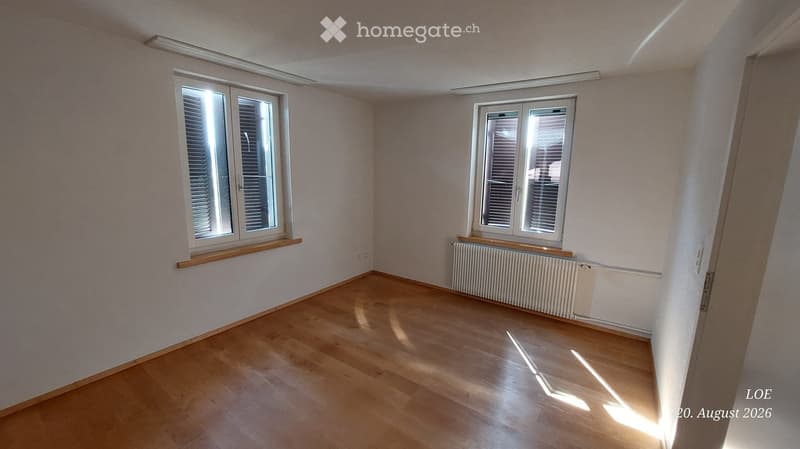 Empty room with wooden flooring, light-colored walls, and two windows with blinds. Radiator visible under one window.