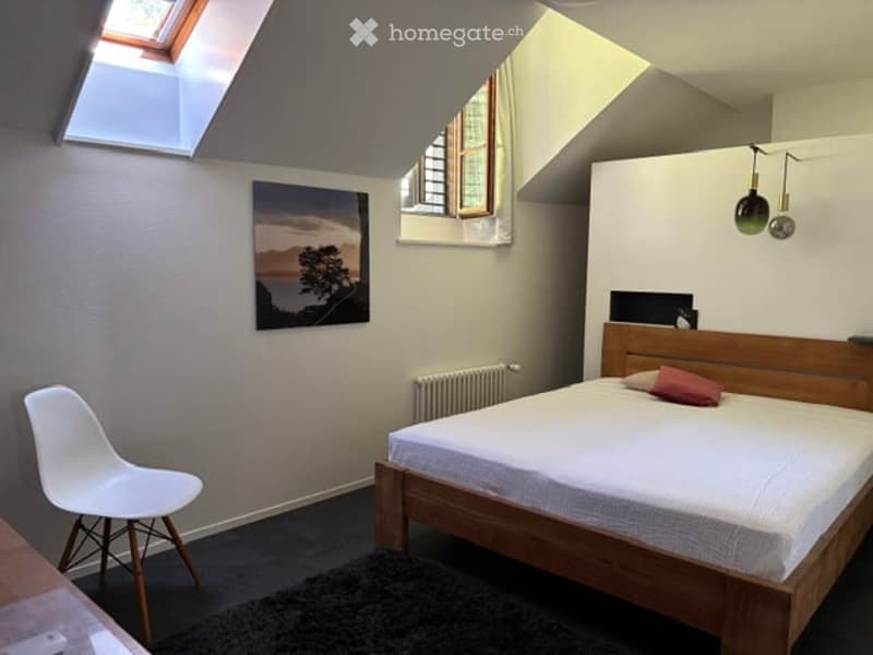Cozy bedroom with a wooden bed, white chair, and a scenic picture on the wall. The room features large windows and a skylight, creating a bright and airy atmosphere.