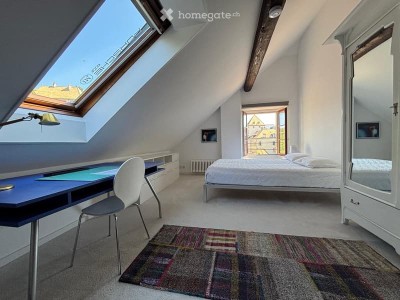 Bright attic bedroom with slanted ceilings, a bed, a desk with a chair, a mirror, and a colorful rug.