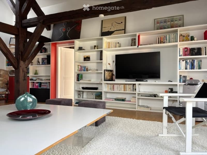 Modern living room with exposed wooden beams, white built-in bookshelves, a flat-screen TV, a white coffee table, and colorful chairs.