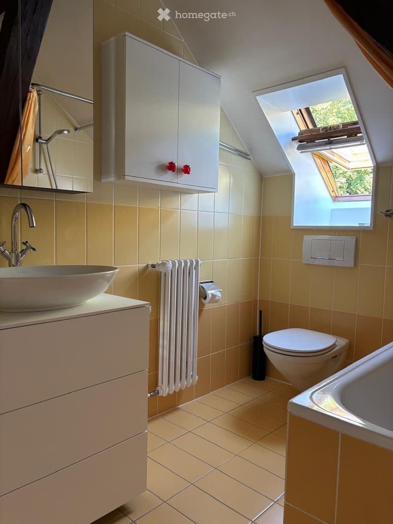 Modern bathroom with beige tiles, a sink, toilet, bathtub, and a skylight window. Features a white cabinet, a mirror, and a radiator with towels.
