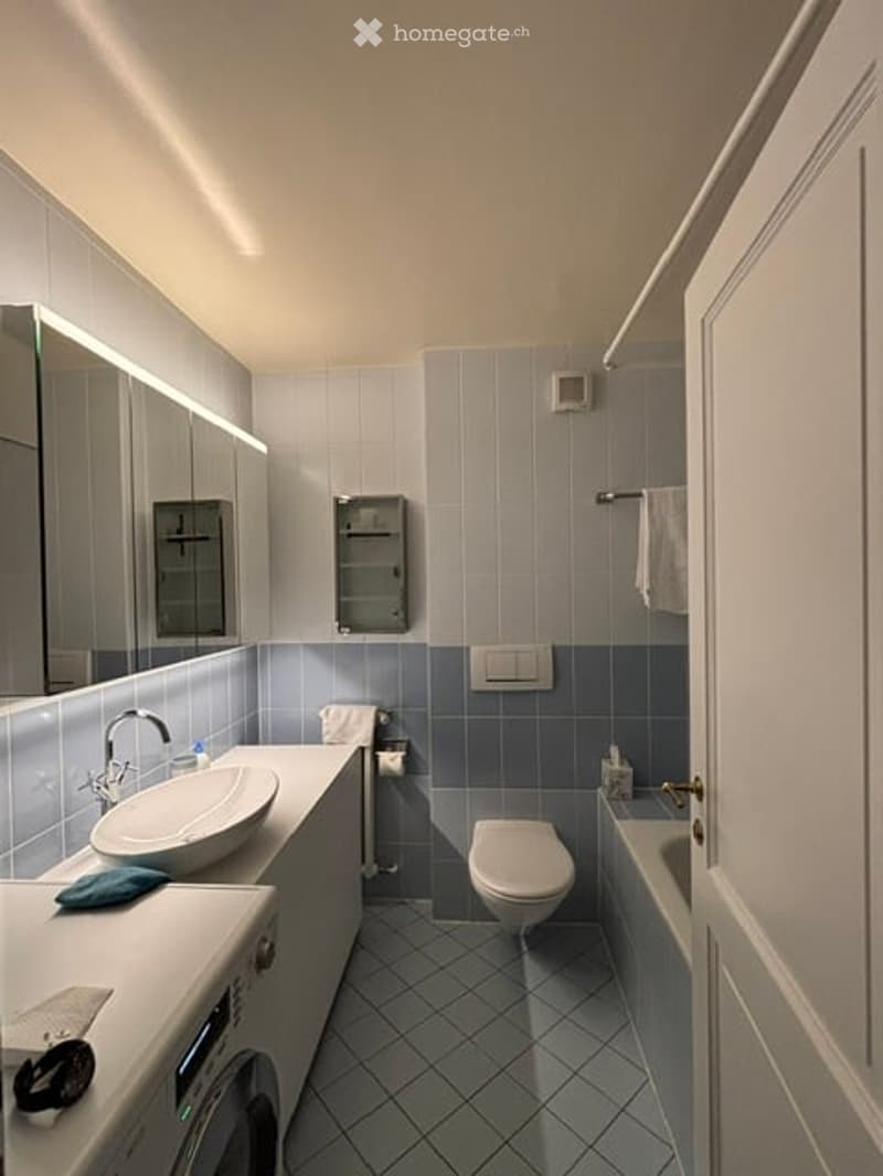Modern bathroom with blue tiles, white sink, mirror, toilet, bathtub, and towel. Features a sleek design with integrated lighting above the mirror.