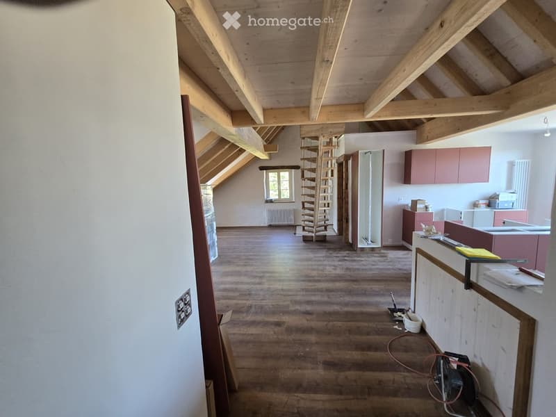 An attic space with exposed wooden beams, wooden flooring, and a small kitchen area with cabinets and a sink. A wooden stool is placed near the kitchen area, and a window provides natural light.
