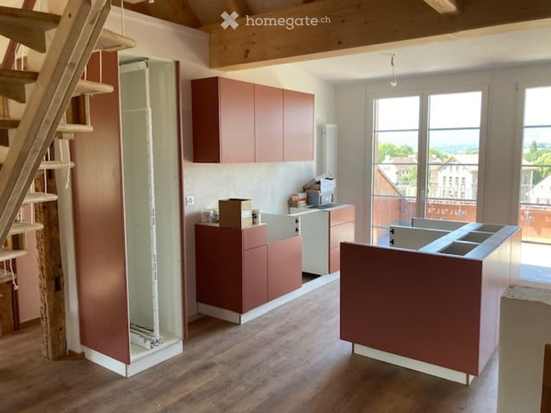 Modern kitchen with wooden flooring, brown cabinets, and a large island. The space features a sink, stove, and fridge, with large windows offering a scenic view.
