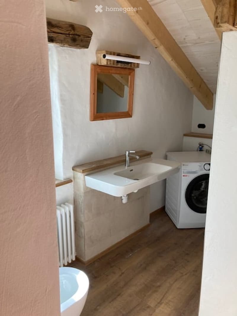 Cozy attic bathroom with wooden beams, a sink, mirror, radiator, washing machine, and toilet.