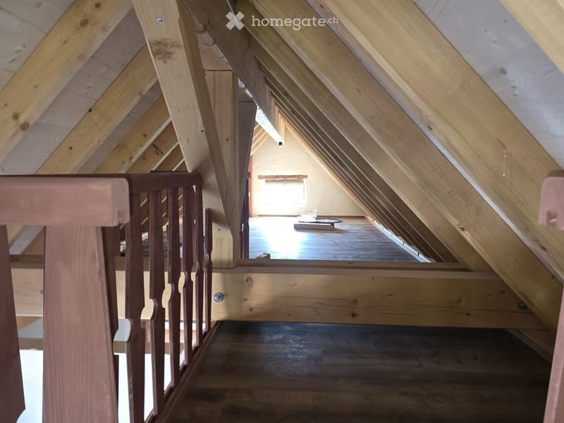 View of an attic space with wooden beams, exposed rafters, and a small window letting in natural light. The floor is wooden, and a wooden railing is visible in the foreground.