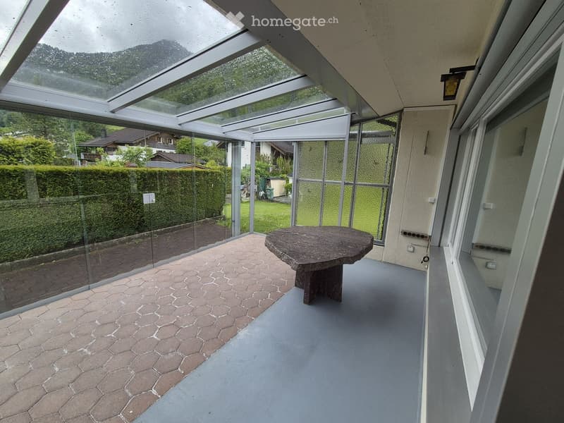 Glass-enclosed balcony with a round stone table, hexagonal tiled floor, and views of a garden and surrounding houses.