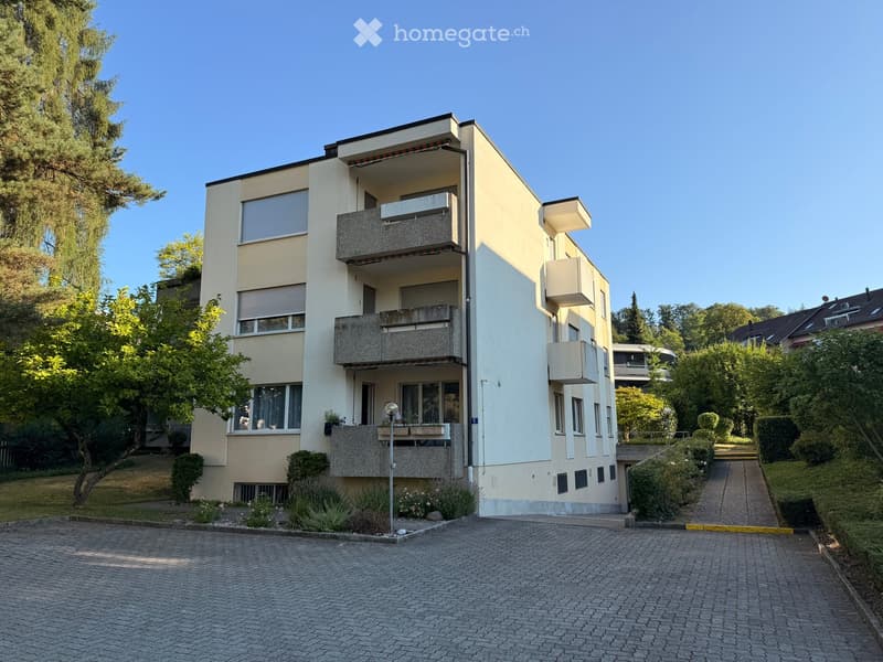 Modern multi-story residential building with balconies, surrounded by greenery and a paved driveway.