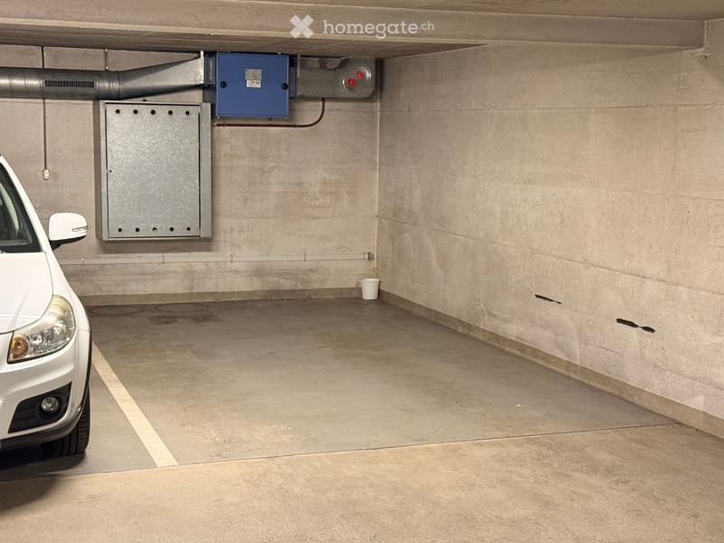 Underground parking space with a single white car parked, concrete walls, electrical panels, and a small white bucket on the floor.