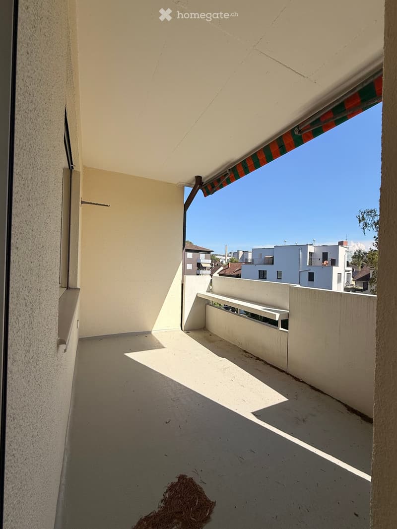 Balcony with a retractable awning, white walls, and a view of neighboring buildings under a clear blue sky.