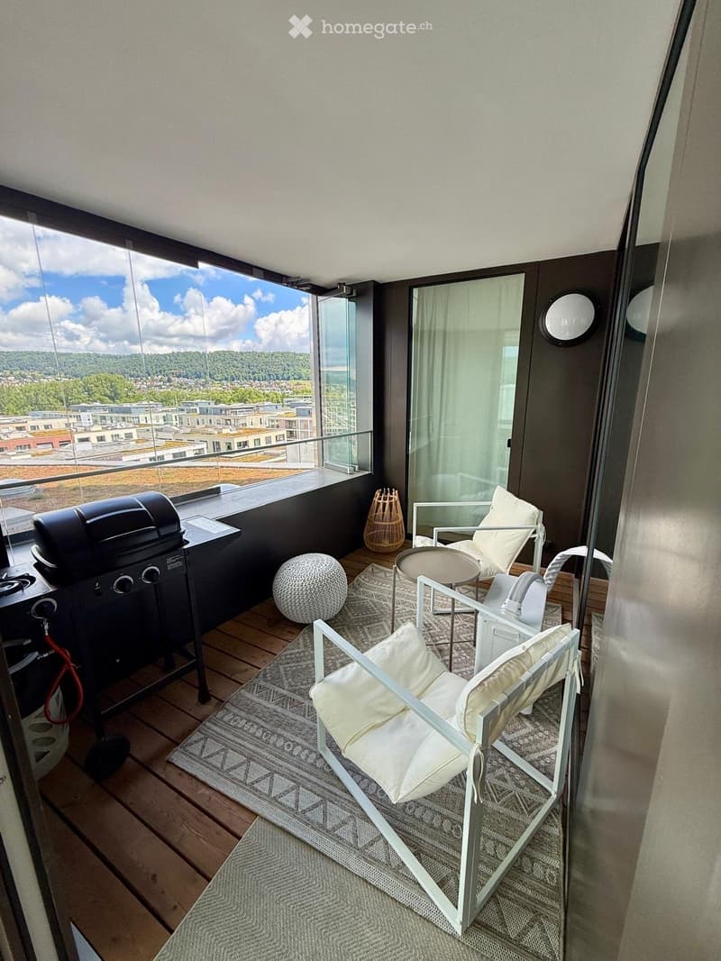 Balcony with wooden decking, modern white chairs, small table, round ball, wicker basket, and grill, overlooking a cityscape with greenery.