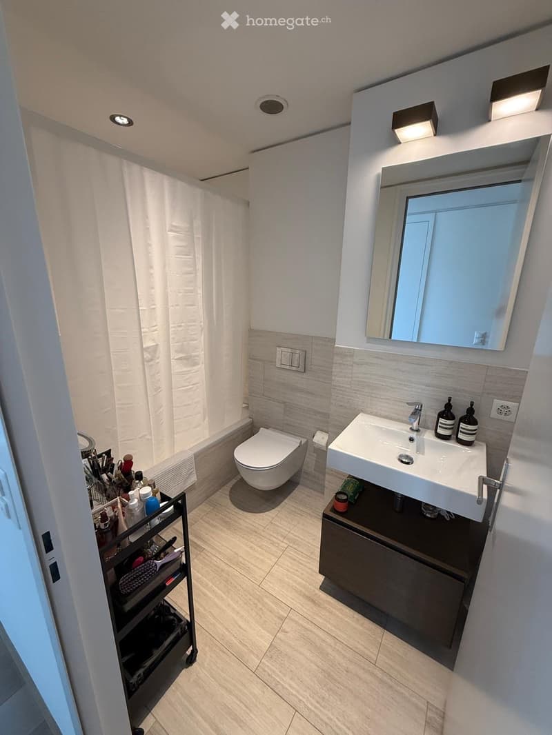 Modern bathroom with a white sink, toilet, bathtub, and mirror. Includes a dark wooden cabinet under the sink and a towel rack with toiletries.