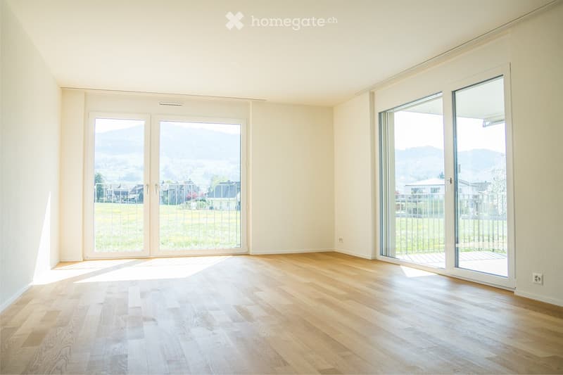 Empty room with wooden flooring, large sliding glass doors, and a scenic view of a green field and distant houses.