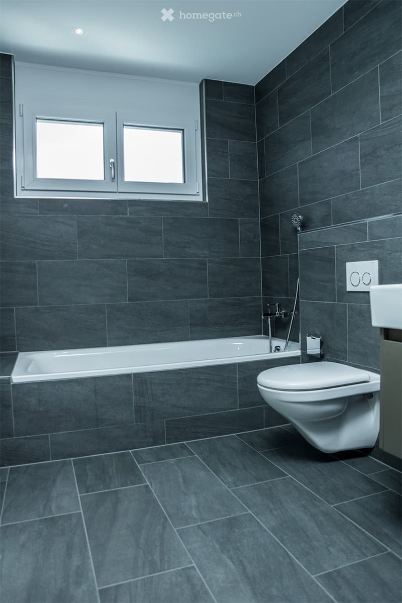Modern bathroom with dark grey tiles, a bathtub, toilet, and a window with two panes.