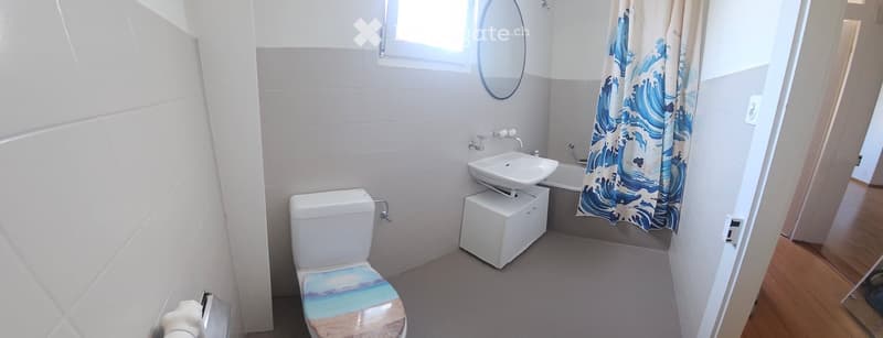 Modern bathroom with white tiles, a toilet with a beach-themed seat cover, a white sink on a cabinet, a round mirror, and a bathtub with a blue ocean-themed shower curtain.