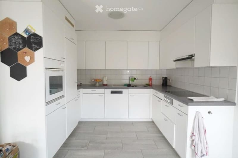 Modern, minimalist kitchen with white cabinets, stainless steel appliances, and a grey countertop. Features an oven, dishwasher, and a sink with a faucet. A towel hangs on the side.