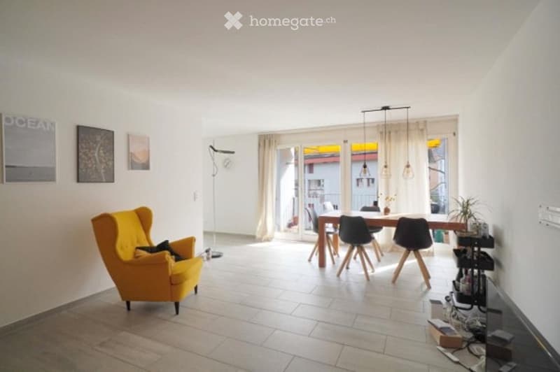 Bright, open living and dining area with white walls, tiled floor, large windows leading to a balcony, yellow armchair, dining table with chairs, floor lamp, and decorative picture frames.