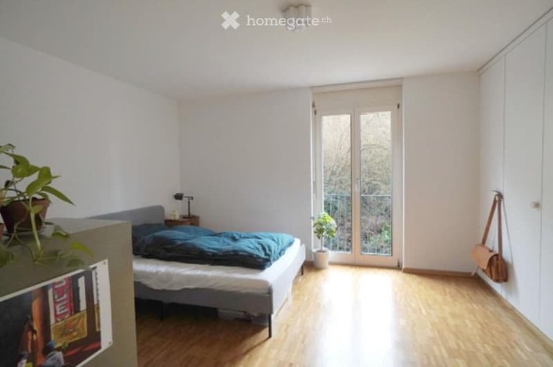 Bright bedroom with wooden flooring, a grey bed with blue bedding, a nightstand with a lamp, and a potted plant near the window. A sliding glass door leads to a balcony.