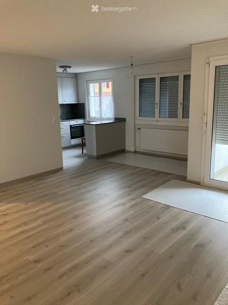 Empty room with wooden flooring, large window, and a view of an adjacent kitchen area with cabinets and appliances.