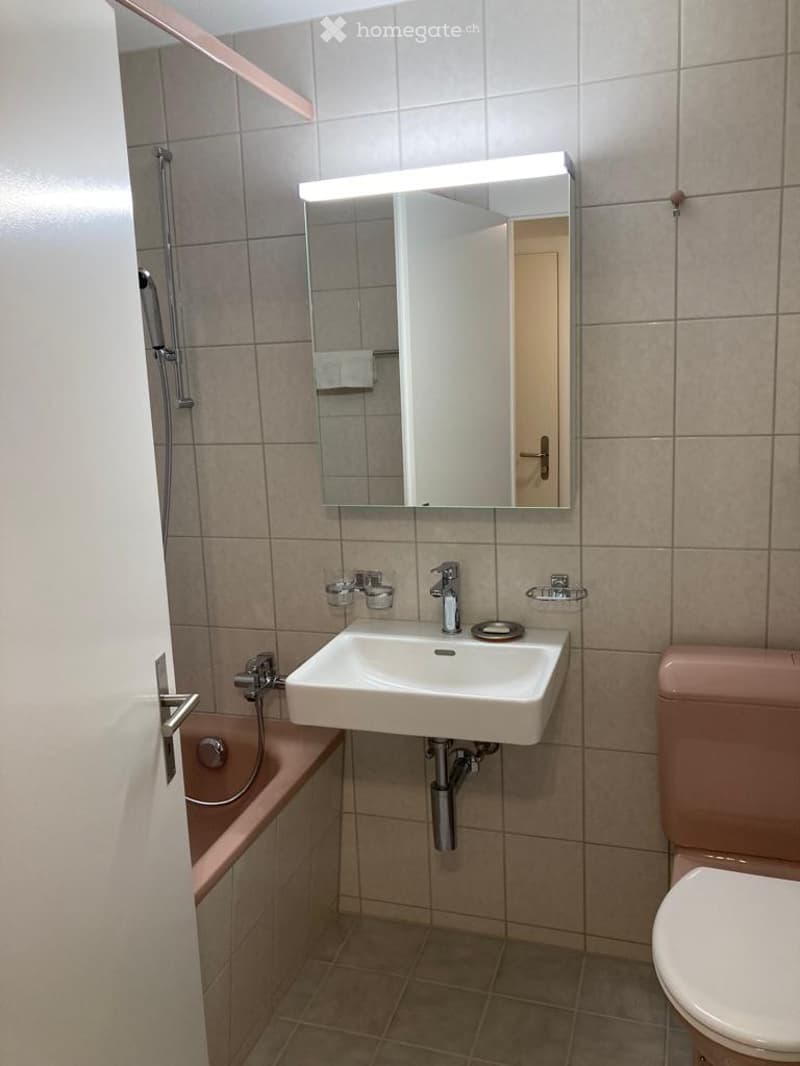 Bathroom with beige tiled walls and floor, white sink with chrome faucet, illuminated mirror, and a pink toilet. A bathtub is visible on the left side.