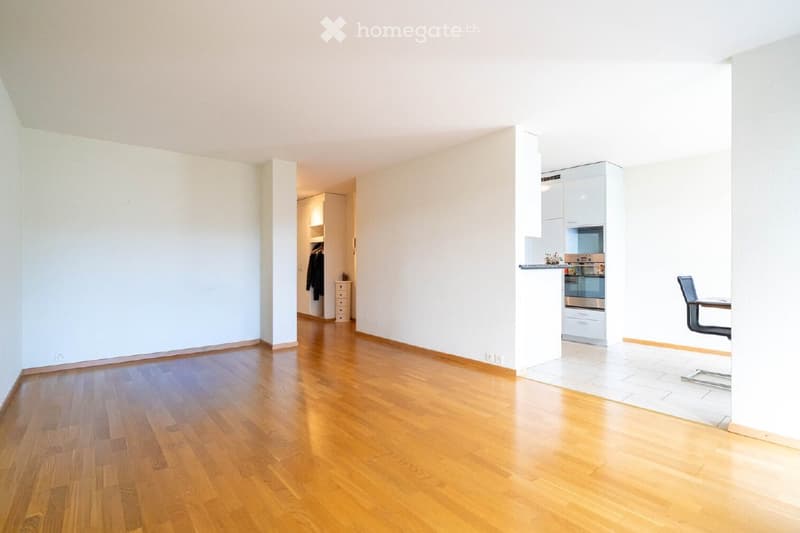 Bright, unfurnished room with wooden flooring, white walls, and a small kitchen area in the corner. The space includes a doorway leading to another room with built-in storage.