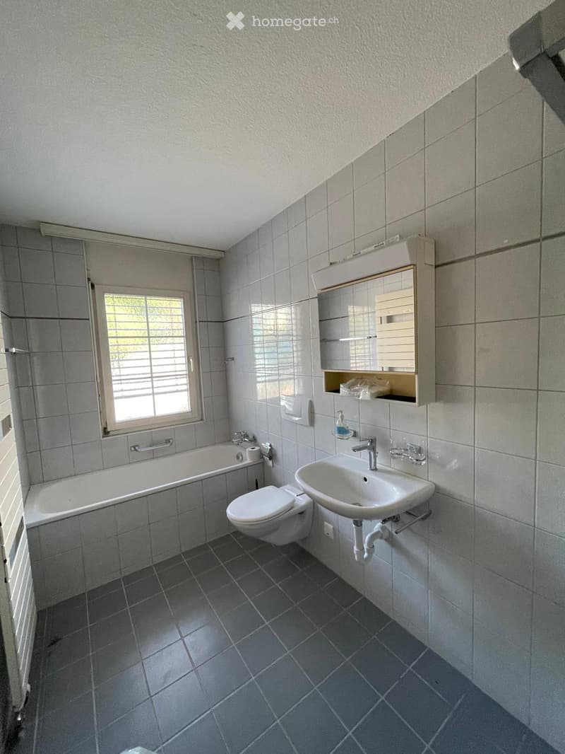 Modern bathroom with grey tiles, bathtub, toilet, sink, mirror, and cabinet.