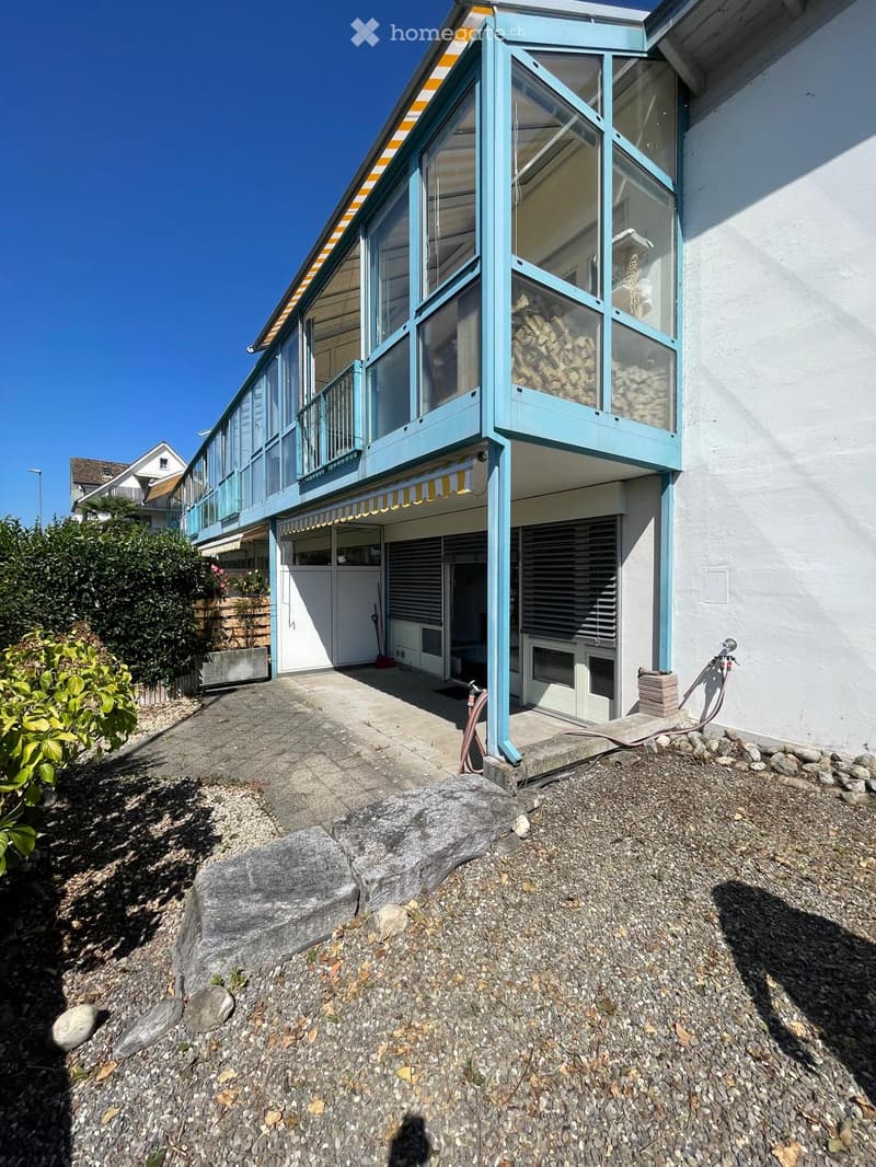 Modern two-story building with a light blue facade, large glass windows, and a covered entrance area. The ground level features a paved area with a small garden and a stone pathway leading to the entrance.