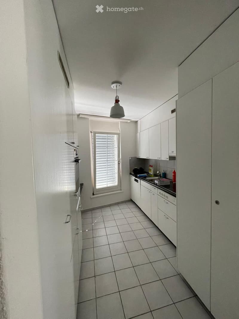 Modern kitchen with white cabinets, tiled floor, and stainless steel appliances including a stove, oven, and fridge. A window with blinds provides natural light.