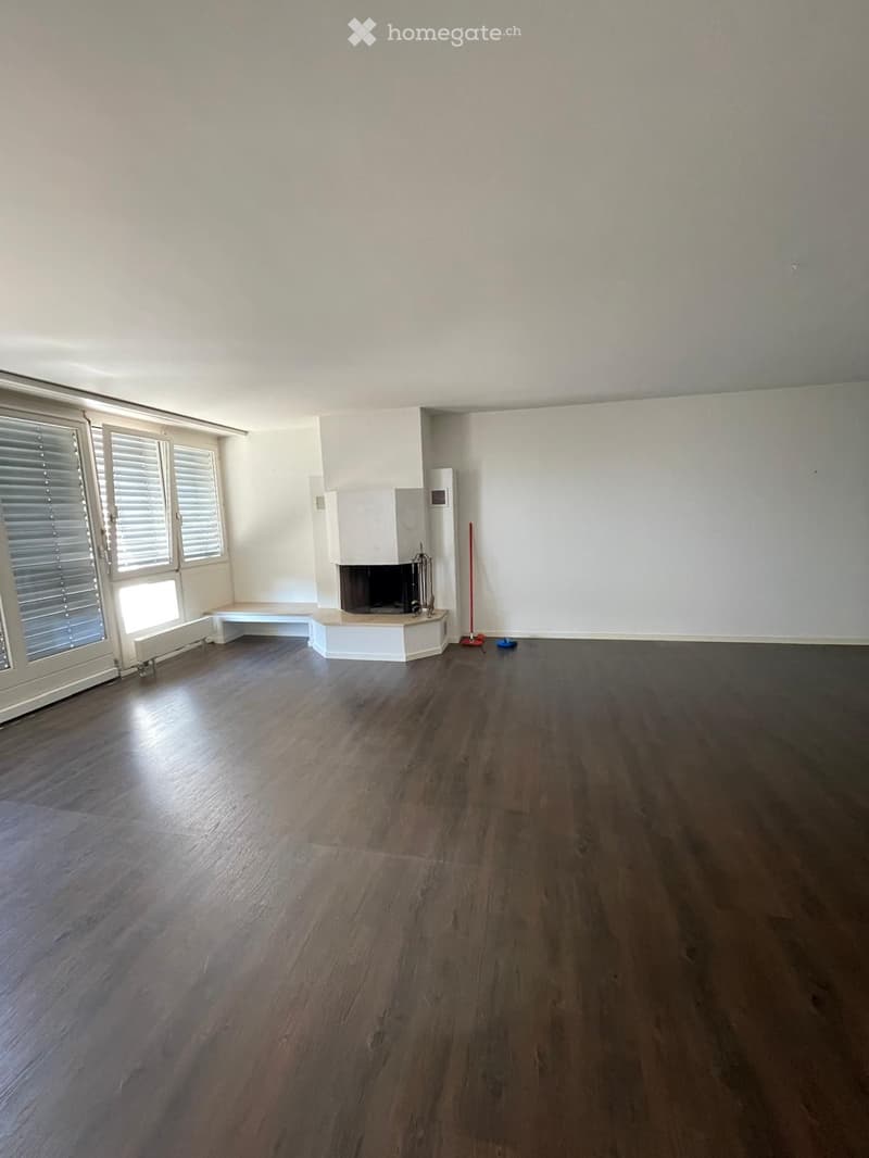 Empty room with dark wooden flooring, white walls, and a fireplace with a bench nearby. Windows with blinds and a radiator are visible.