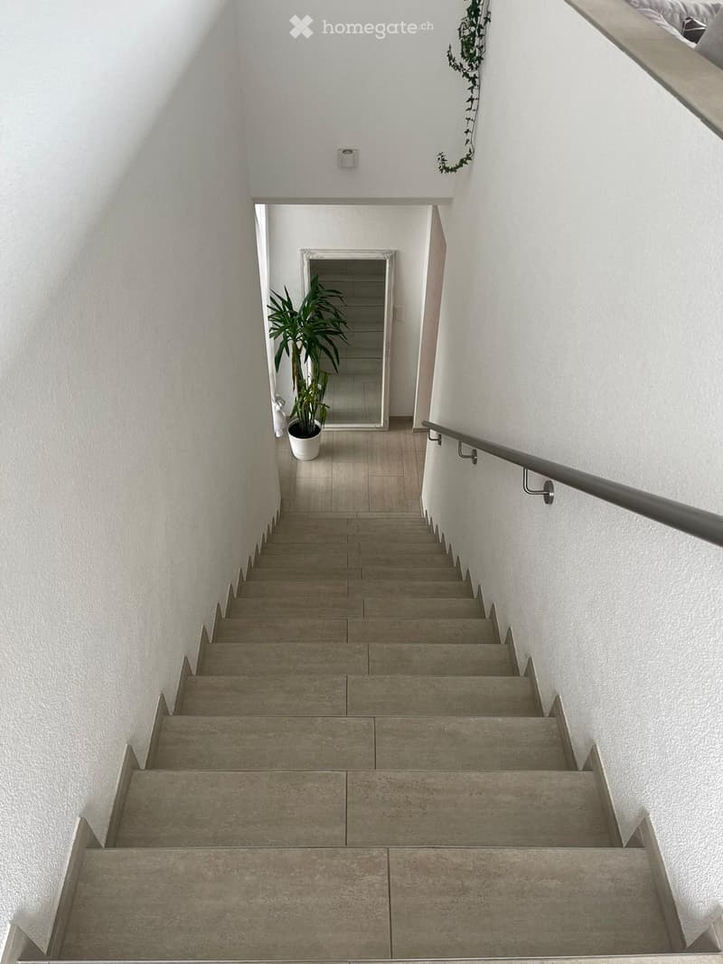 Staircase with beige tiled steps, white walls, a metal handrail, and a potted plant in the background.