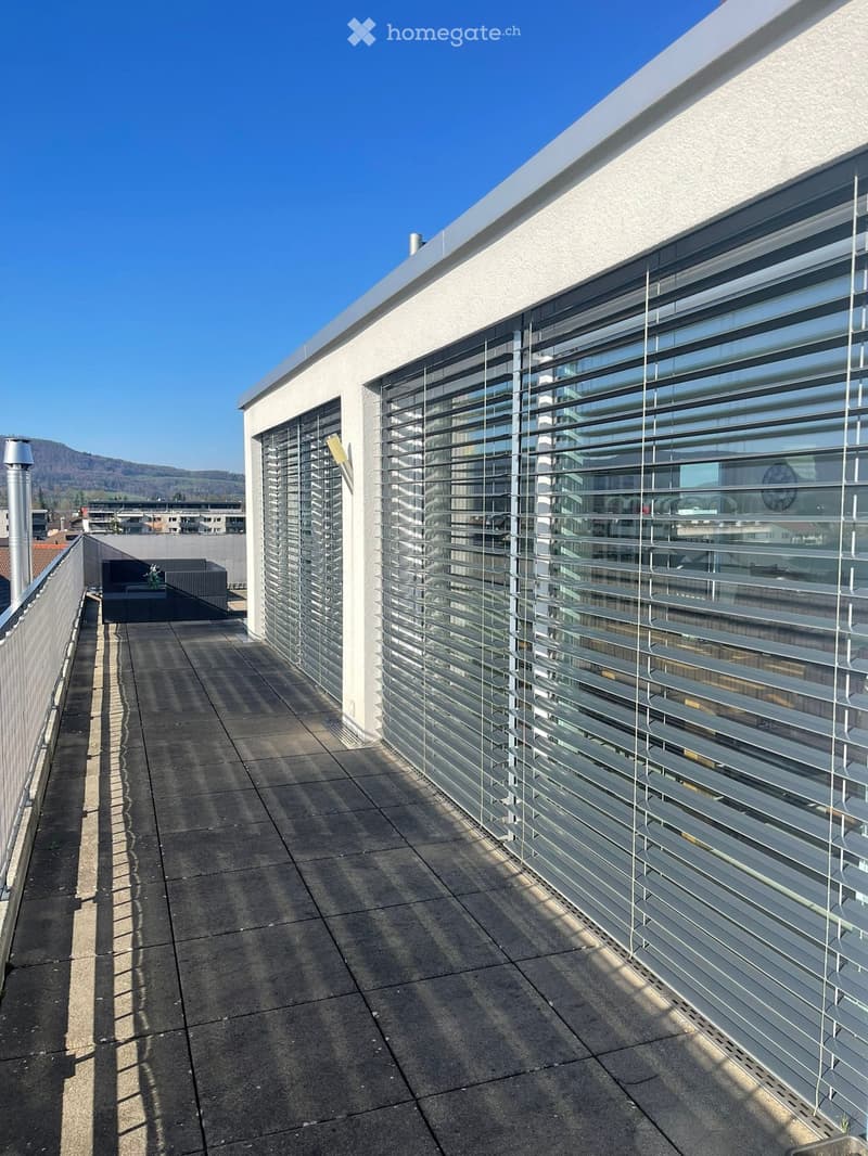 Modern rooftop terrace with large glass windows and blinds, concrete floor, and a small seating area with a sofa and table.