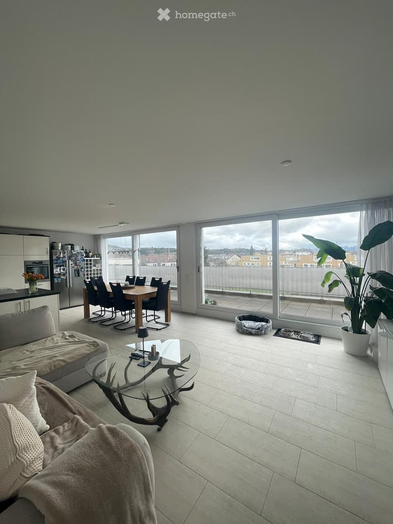 Open-plan living and dining area with large windows, a sofa, coffee table, dining table with chairs, and a kitchen area with appliances.