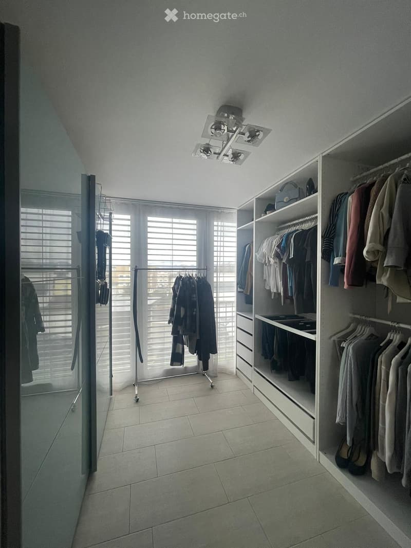 Modern walk-in closet with built-in wardrobe, clothing rack, and storage shelves. Features tiled flooring, large windows with blinds, and multiple hanging clothes.