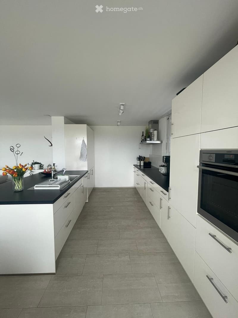 Modern kitchen with white cabinets, dark countertops, and stainless steel appliances. Features a sink, oven, coffee maker, and decorative vase with flowers.