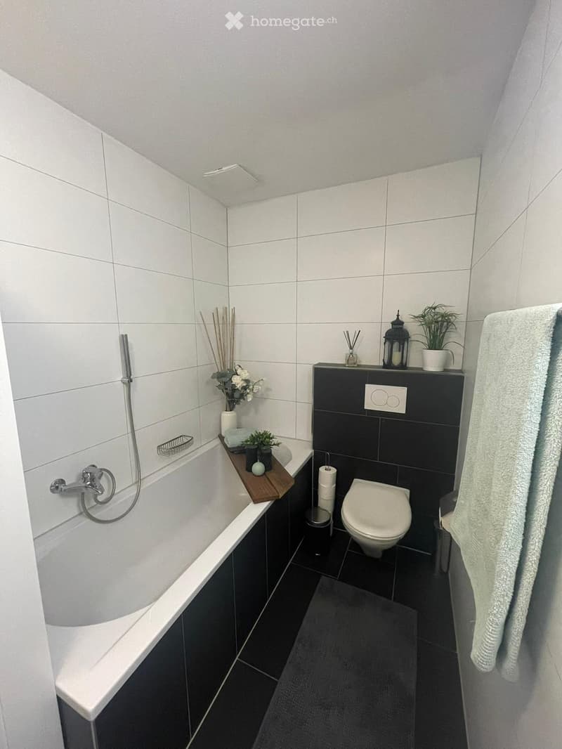 Modern bathroom with white tiles, a bathtub, toilet, towel, and decorative items including plants and candles.