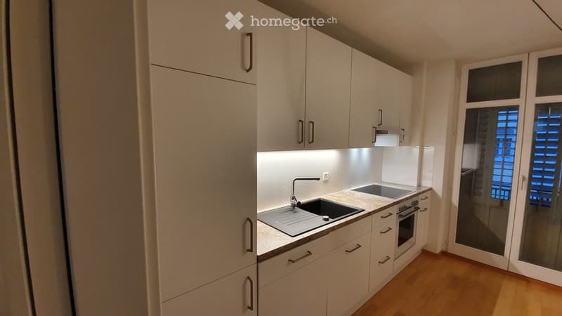 Modern kitchen with white cabinets, a sink, and an oven, featuring a wooden floor and a window with blinds.