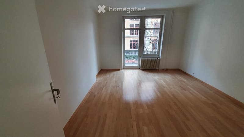 Empty room with wooden flooring, white walls, and a large window with a view of a building and balcony.