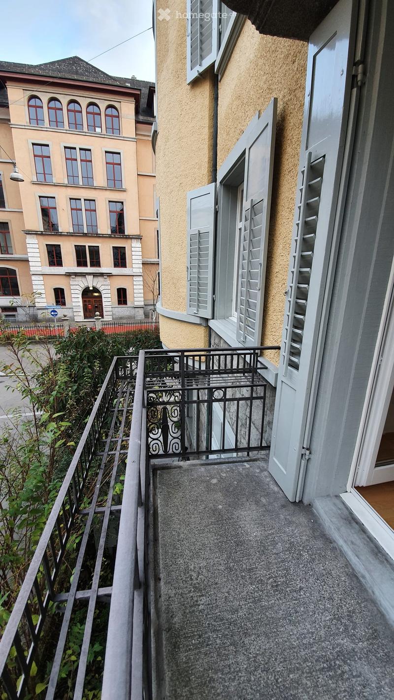 Balcony with grey concrete floor, black metal railing, open shutters, and greenery visible below.